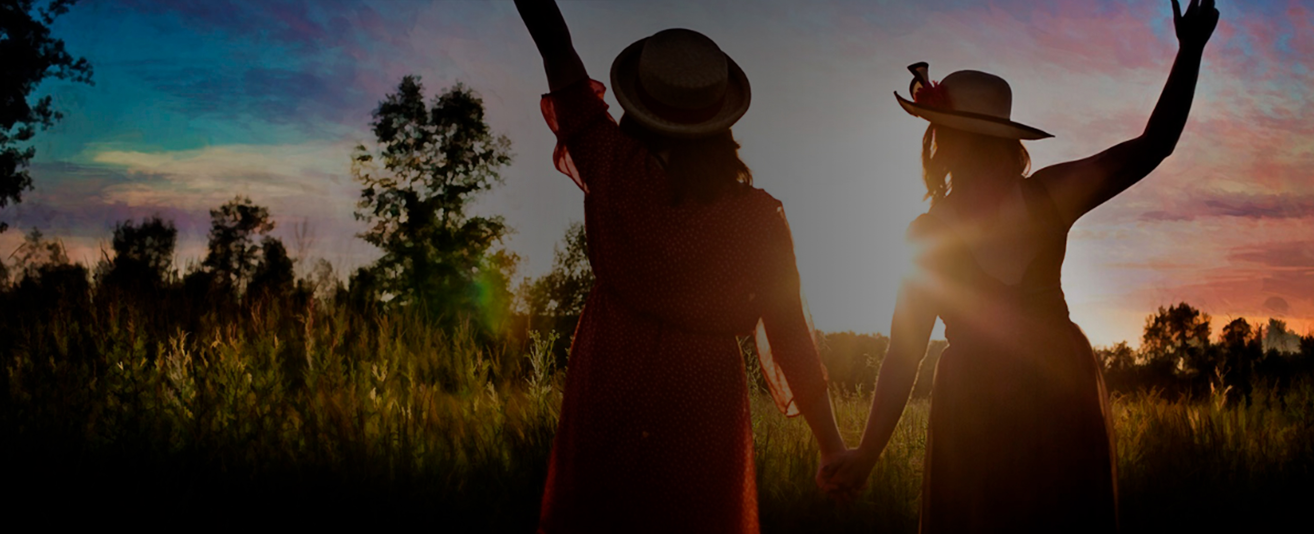 A photograph of two women with there backs to the camera looking into the sun rise with there hands raised in the air.